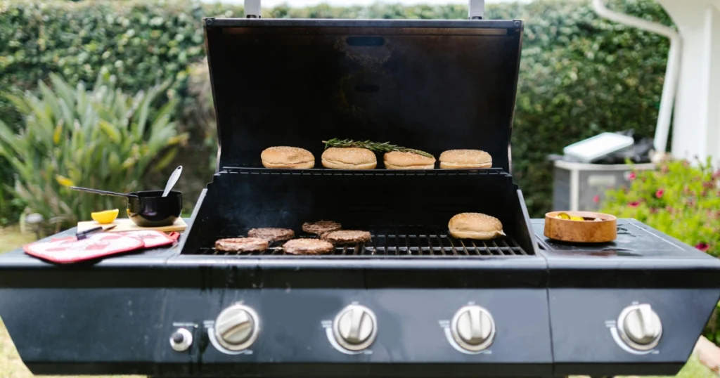 A backyard barbecue grill features burger patties sizzling below and buns with rosemary sprigs placed on the top rack, while cooking utensils and a bowl of ingredients sit beside it, showing what to cook on a griddle.